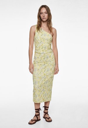 Femme debout portant une robe midi jaune à fleurs, sans manches et à une épaule, et des sandales plates marron à lacets sur un fond blanc.