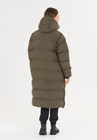Langer, olivgrüner Puffer-Mantel mit Kapuze, der ein horizontales Steppmuster, seitliche Schlitze und gerippte Bündchen aufweist. Kombiniert mit schwarzen Hosen und Stiefeln.