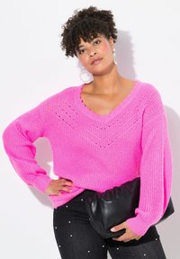 Maglione rosa chiaro a maglia con scollatura a V e dettagli a punto aperto, abbinato a pantaloni neri con accenti di perle e una pochette nera.