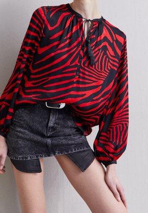 Personne portant un chemisier à manches longues zébré rouge et noir avec des liens à pompons et une mini-jupe en denim noire avec des panneaux triangulaires en cuir.