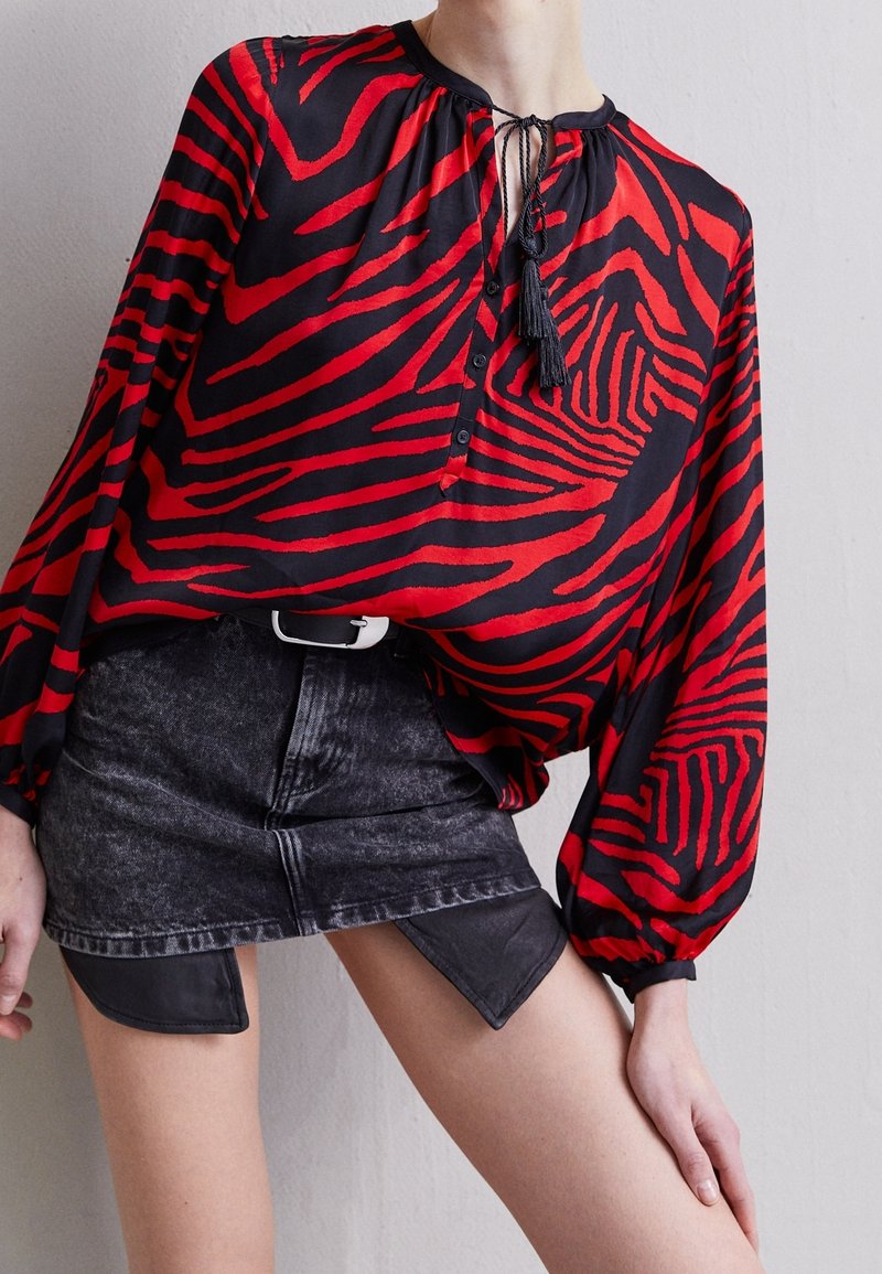 Personne portant un chemisier à manches longues zébré rouge et noir avec des liens à pompons et une mini-jupe en denim noire avec des panneaux triangulaires en cuir.