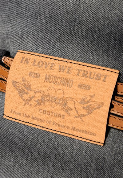 Etiqueta de couro castanho com texto em relevo e desenhos de pássaros, presa a um tecido cinza texturizado, com bordas costuradas e um estilo de etiqueta clássica.