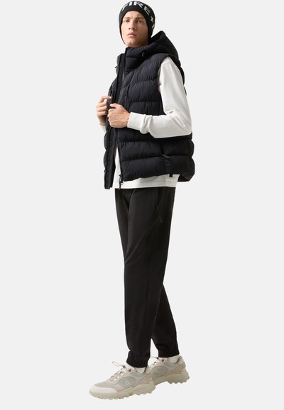 Černá péřová vesta s kapucí, nošená přes bílou dlouhou košili. Černé tepláky a světle šedé tenisky s béžovými akcenty doplňují outfit.