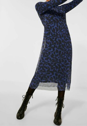 Personne portant une robe midi bleue à motifs transparente sur des leggings noirs, associée à des bottes de combat noires à lacets, posant devant un fond uni.