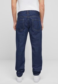 Jeans de mezclilla índigo con un corte recto, costura visible, dos bolsillos traseros y textura sutil. Llevados con una camisa blanca y zapatillas blancas.
