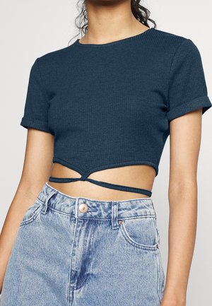 Vrouw draagt een donkerblauw geribd crop top met wikkelbanden en een hooggetailleerde lichtblauwe spijkerbroek.