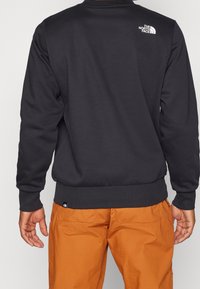 Sudadera negra con puños y dobladillo acanalados, presenta un logo blanco en la parte superior de la espalda, combinada con pantalones cargo naranjas.