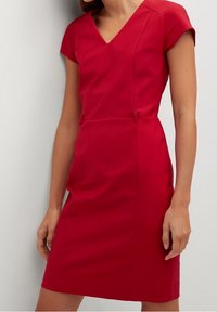 Robe rouge mi-mollet avec une silhouette ajustée, des manches courtes et un décolleté en V. Elle présente de légères fronces sur les côtés et des accents décoratifs à la taille.