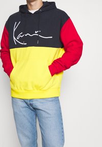 Felpa con cappuccio con sezioni nere, rosse e gialle e un logo bianco ben evidente. Realizzata in tessuto morbido, con tasca frontale.