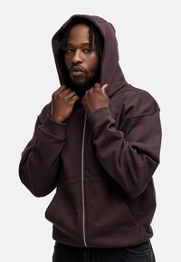 Dunkellila Zip-Hoodie mit großer Kapuze, ausgestattet mit gerippten Bündchen und Saum. Das Material wirkt weich und strukturiert und hat Kängurutaschen.