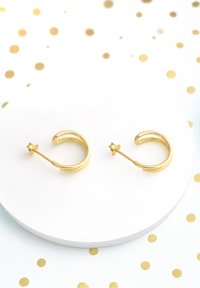 Goldene Creolenohrringe mit einem glatten, abgerundeten Design, ausgestattet mit einem Steckverschluss und einem dezenten dekorativen Akzent. Auf einer weißen Oberfläche präsentiert.