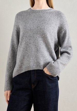Personne portant un pull gris en tricot, légèrement rentré dans un jean bleu foncé, avec une main dans la poche, sur un fond neutre.