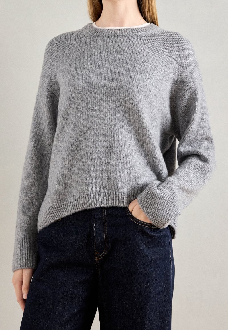 Personne portant un pull gris en tricot, légèrement rentré dans un jean bleu foncé, avec une main dans la poche, sur un fond neutre.