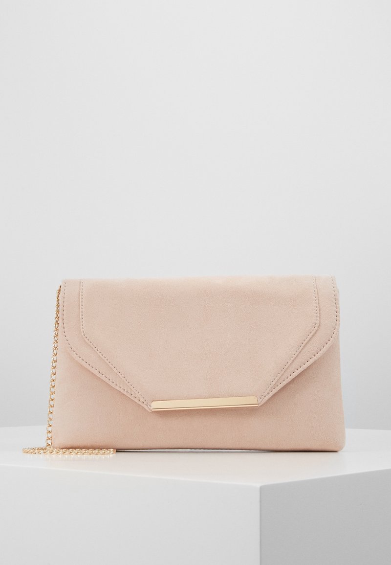 Pochette en daim beige avec un détail barre dorée sur le rabat et une fine bandoulière chaîne dorée posée sur une surface blanche.