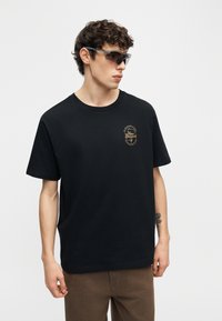 Νέος άνδρας με μαύρο μπλουζάκι New Balance, καφέ παντελόνι και αθλητικά σκουρόχρωμα γυαλιά ηλίου, στέκεται μπροστά σε απλό φωτεινό φόντο.