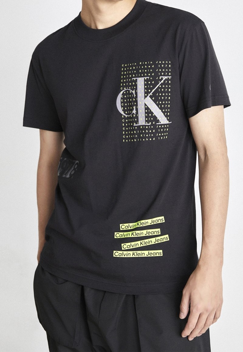 Black cotton T-shirt featuring a graphic print with "CK" in gray and yellow text, and brand labels in yellow on the hem.
