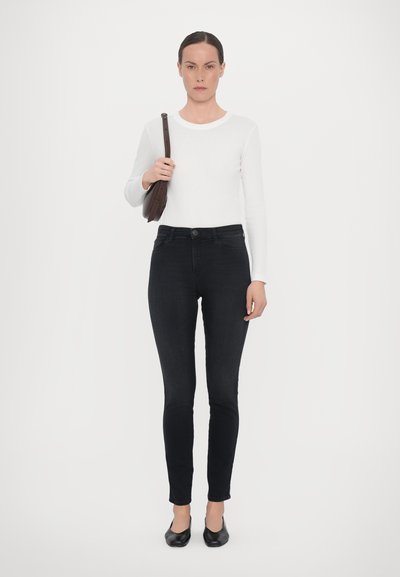 Svarta skinny jeans i kombination med en långärmad vit topp. Modellen håller en brun axelväska. Outfiten har en figurnära design och enkla texturer.