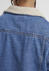 Denim jacket with a light blue wash, featuring a cream fleece collar. Visible stitching and a relaxed fit frame the back view.