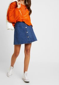 Pomarańczowy sweter dziany z detalem z otwartym splotem oraz ciemnoniebieska spódnica w linii A z pięcioma srebrnymi guzikami z przodu. Białe sneakersy z wysoką cholewką.