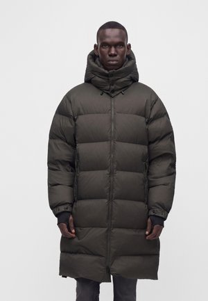 Homme portant un long manteau d’hiver matelassé vert foncé avec un col montant et une capuche, debout devant un fond clair uni.