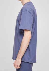 Uomo che indossa una semplice t-shirt viola oversize a maniche corte e pantaloni coordinati, in posa di profilo su uno sfondo grigio chiaro.