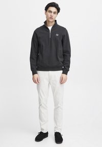 Zwarte half-zip sweatshirt met een klein logo, gecombineerd met witte slim-fit broeken en zwarte suède schoenen. Strak ontwerp en aansluitende vorm.