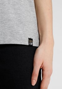 Camiseta gris con una textura suave, que presenta una etiqueta negra con "SUPER DRY" en el dobladillo. Mano reposando sobre una tela negra.