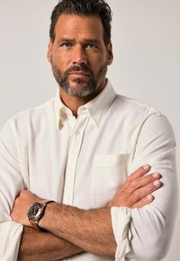 Witte overhemd met knopen en een enkele borstzak, textuurstof en opgerolde mouwen. Pols van de drager draagt een zilveren horloge met een zwarte wijzerplaat.