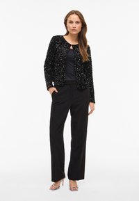 Črna jakna s sequini, krogu srčnega izreza in dolgimi rokavi; v kombinaciji z črnimi, ohlapnimi hlačami in kovinskimi sandalmi s peto.