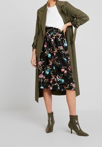 Olijfgroene trenchcoat over een witte t-shirt, gecombineerd met een zwarte bloemenrok met kleurrijke patronen en groene block-ankelschoenen.