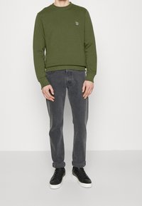 Felpa verde oliva con scollo tondo, abbinata a jeans grigi e sneaker nere. Presenta un piccolo dettaglio ricamato sul petto.