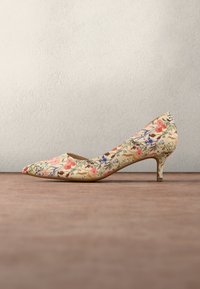 Escarpin beige à talon bas avec motif floral et bout pointu, reposant sur une surface en bois face à un mur clair uni.