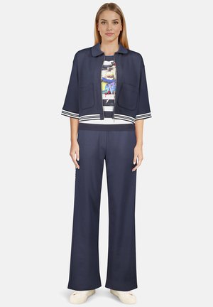 Veste courte bleu marine avec des rayures blanches, manches courtes, et deux poches avant, associée à un pantalon à jambes larges et un T-shirt graphique multicolore.