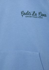 Tissu bleu clair avec texte cursif vert foncé « Goditi La Cena » et lettres en bloc « Savor Your Food » au-dessus d'une couture de poche kangourou.