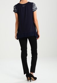 Kvinna i marinblå topp med mönstrade ärmar, svarta slim fit byxor och svarta högklackade skor, stående vänd bort från kameran mot vit bakgrund.