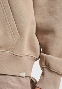 Sudadera beige con bolsillo canguro, puños y dobladillo acanalados, y una pequeña etiqueta de marca en la costura inferior. Textura suave de mezcla de algodón.