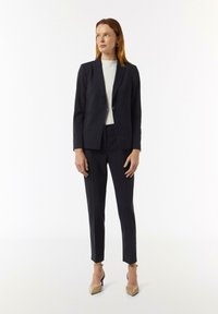 Dunkel gestreifter Anzug mit einem taillierten Blazer und schmal geschnittenen Hosen, kombiniert mit einem hellen Oberteil und spitzen Stilettos.