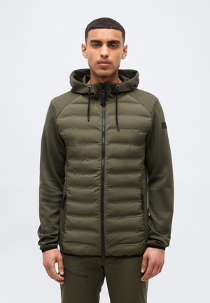 Férfi olívazöld steppelt, cipzáras kapucnis dzsekiben, húzózsinórokkal, egy egyszínű világosszürke háttér előtt áll.