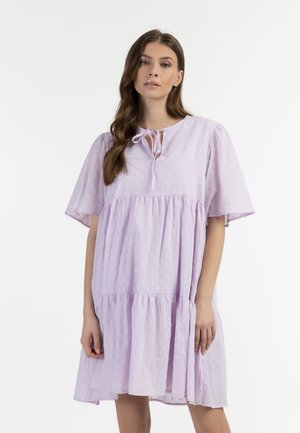 Junge Frau mit langen braunen Haaren, die ein lockeres, hellviolettes Kleid mit kurzen Ärmeln und einem Bindeband am Ausschnitt vor einem schlichten weißen Hintergrund trägt.