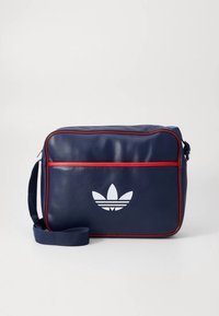 Mala de ombro de pele sintética azul-marinho com piping vermelho, apresentando um logótipo branco trevo na frente e uma alça longa ajustável.