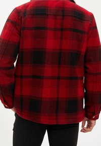 Close-up van een persoon die een rood en zwart geruit flanellen shirt en zwarte broek draagt, gezien van achteren.