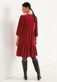 Rotes Samtkleid mit mehrlagigem Rüschensaum, Dreiviertelärmeln und rundem Ausschnitt. Getragen mit kniehohen schwarzen Stiefeln.