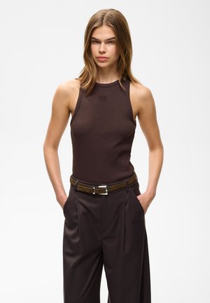 Jeune femme aux cheveux bruns clairs et raides porte un haut sans manches marron foncé, un pantalon taille haute foncé et une ceinture marron avec une boucle argentée.