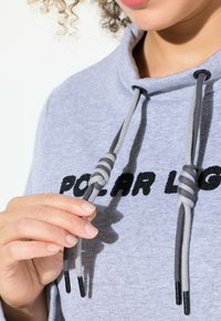 Sudadera gris con un alto cuello, que presenta texto bordado en negro. Los cordones son de dos tonos, gris y negro con puntas de metal negras.