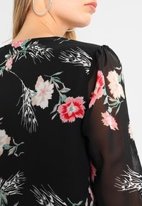 Donna che indossa una blusa nera trasparente con stampa floreale rosa e bianca, visibile dal collo alla parte superiore della schiena e del braccio, con orecchino a cerchio argentato.