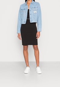 Ljusblå kort denimjacka över en svart figursydd klänning, kombinerad med vita sneakers. Jackan har två bröstfickor och en logotyp.