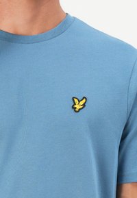 Tricou din bumbac albastru, cu guler rotund și un mic logo brodat în formă de păsărele galbenă pe pieptul stâng.