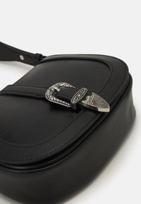 Černá kožená crossbody taška se stříbrnou dekorativní přezkou, s hladkým povrchem a obdélníkovým tvarem s zaoblenými okraji.