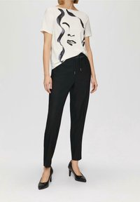 Kvinna iklädd vit t-shirt med tryck av svart abstrakt ansikte, svarta skräddarsydda byxor med dragsko i midjan, och svarta högklackade skor som står mot en enfärgad bakgrund.