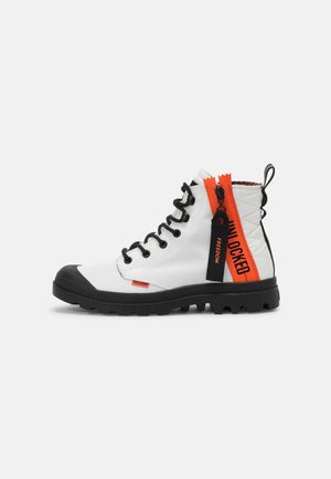 Botte montante blanche et noire avec une semelle épaisse, des lacets noirs et des tirettes de fermeture éclair orange vif étiquetées "UNLOCKED" et "FREEDOM."